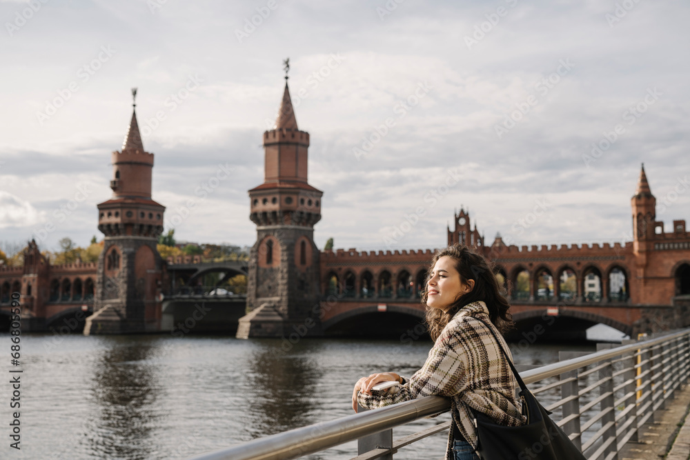 Fototapeta premium Smiling woman with smartphone in the city at Oberbaum Bridge, Berlin, Germany