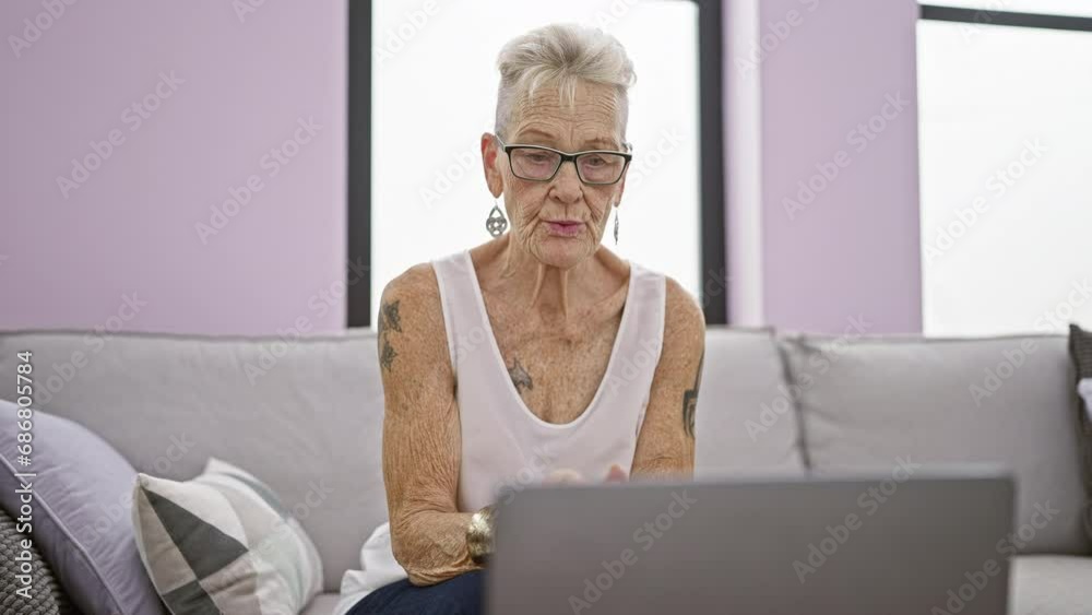 Focused elderly woman with grey hair and glasses, using laptop with concentration while sitting on sofa indoors at home, embodying mature technology use in the living room