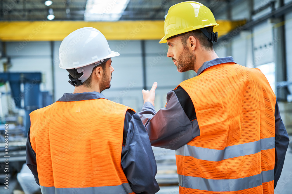Rear view of two men wearing protective workwear talking in factory Rear view of two men wearing protective workwear talking in factory