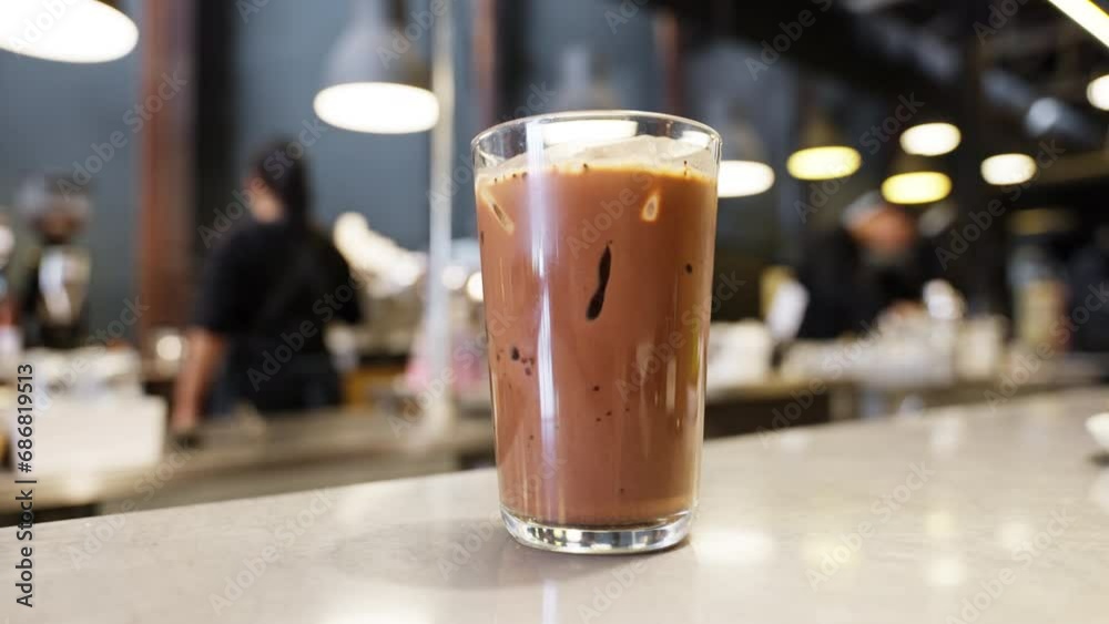 Indulgence awaits in a cafe setting with a tempting iced chocolate placed. The cool drink beckons against a backdrop of a bustling cafe, where a barista walks, adding an energetic touch to the scene.