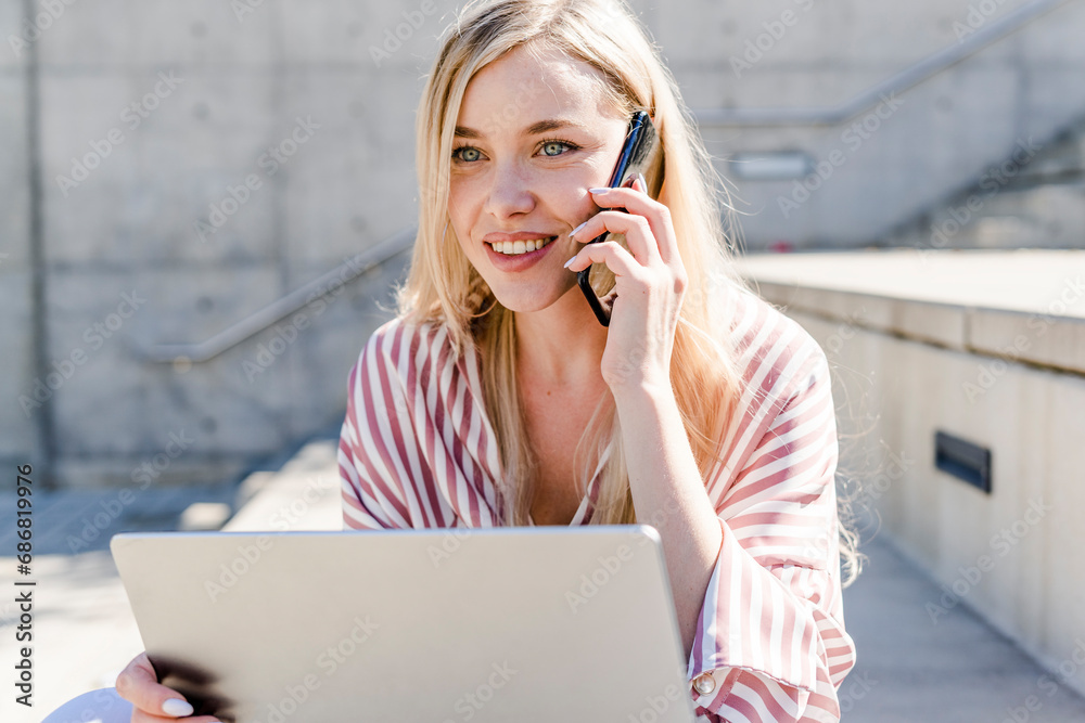 Portrait of smiling blond young woman with laptop on the phone outdoors
