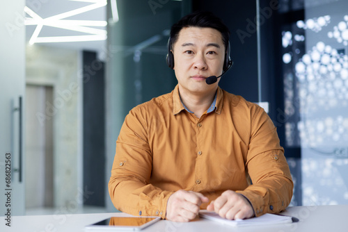 Fotografija Portrait of a young Asian male businessman sitting at a desk in an office wearing a headset and looking seriously at the camera