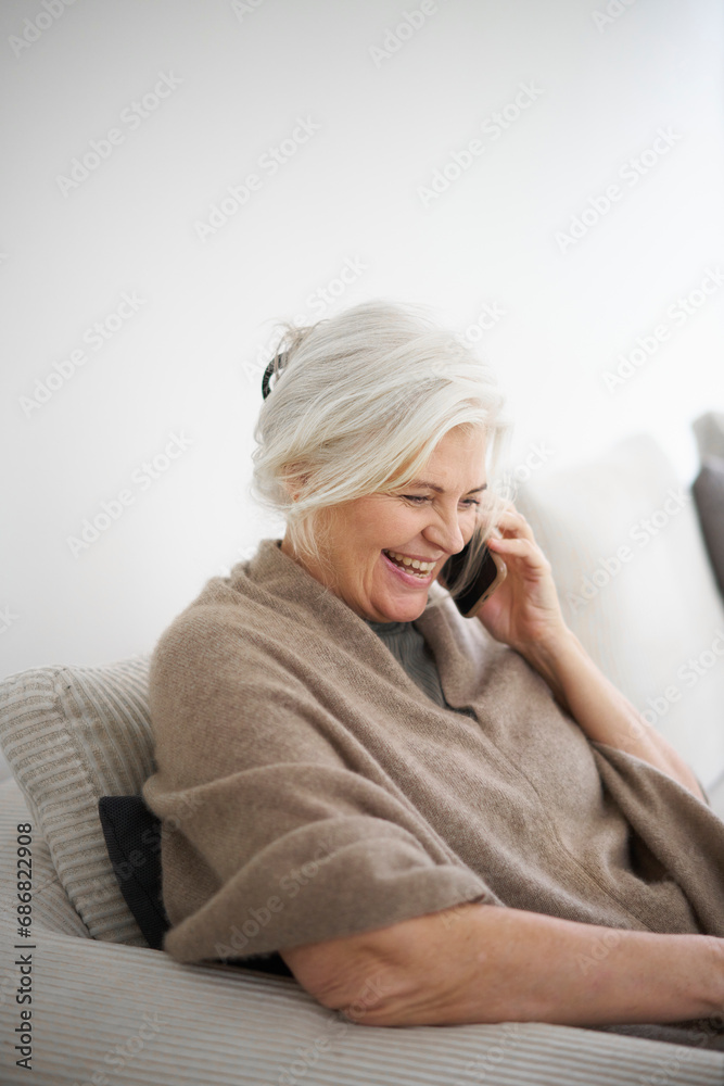 Cheerful senior woman talking on mobile phone while sitting against white wall at home Cheerful senior woman talking on mobile phone while sitting against white wall at home