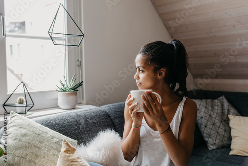 Young woman with cup of coffee sitting on the couch at home looking out of window photo