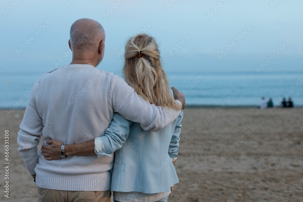 Spain, Barcelona, rear view of senior couple embracing on the beach at dusk
