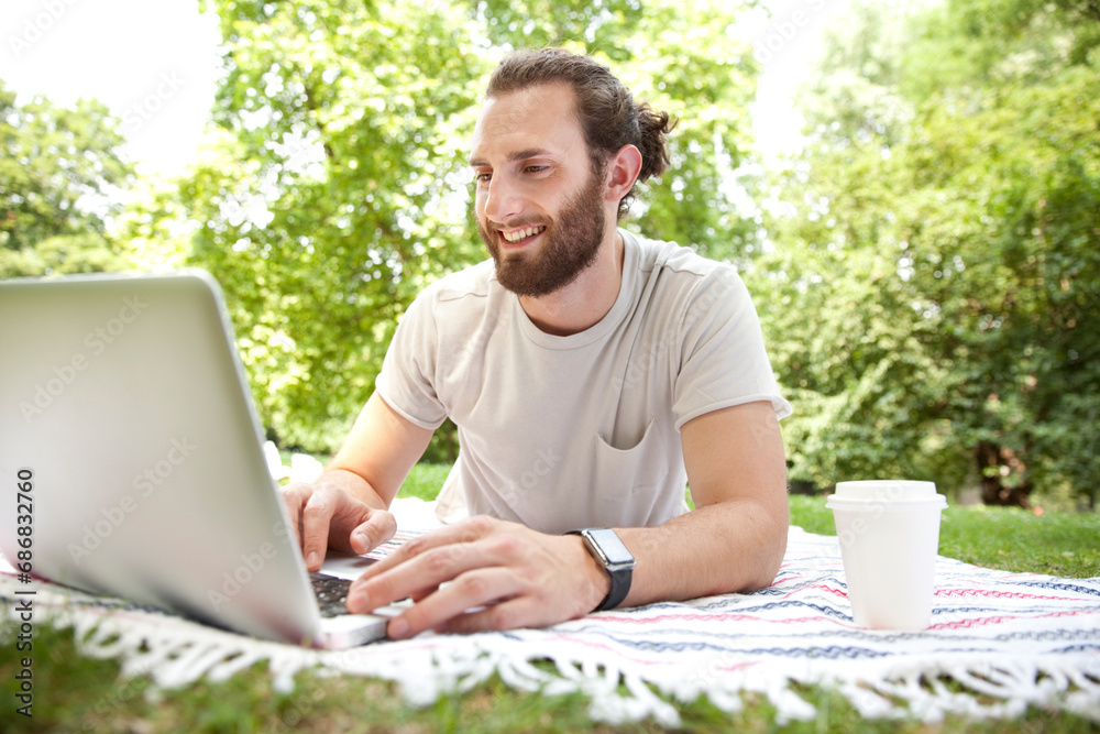 Smiling man with coffee to go lying on blanket in a park using laptop Smiling man with coffee to go lying on blanket in a park using laptop