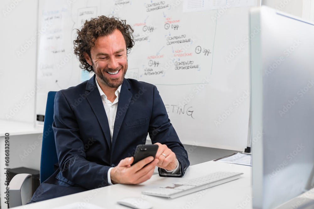 Smiling businessman sitting at desk in office using cell phone Smiling businessman sitting at desk in office using cell phone