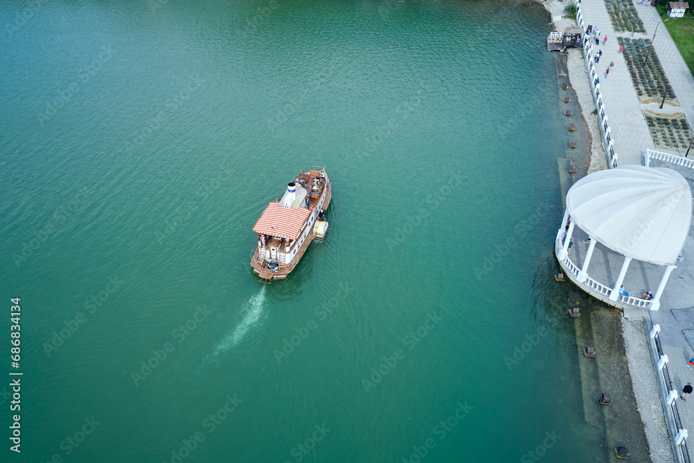 A lone retro-style excursion ship sails along the emerald water surface to the pier. Shot from a drone.
