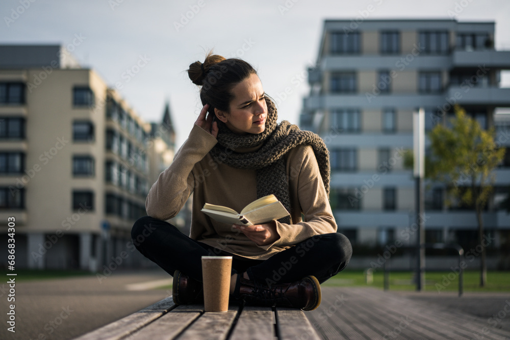 Melancholy woman with coffee to go and book sitting on bench in autumn