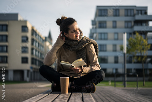 Melancholy woman with coffee to go and book sitting on bench in autumn