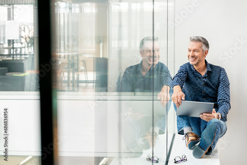 casual businessman sitting on windowsill in office building, using laptp photo