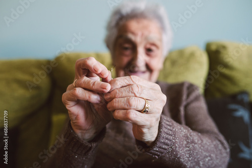 Hands of senior woman sitting on couch passing thread through buttonhole of sewing needle photo
