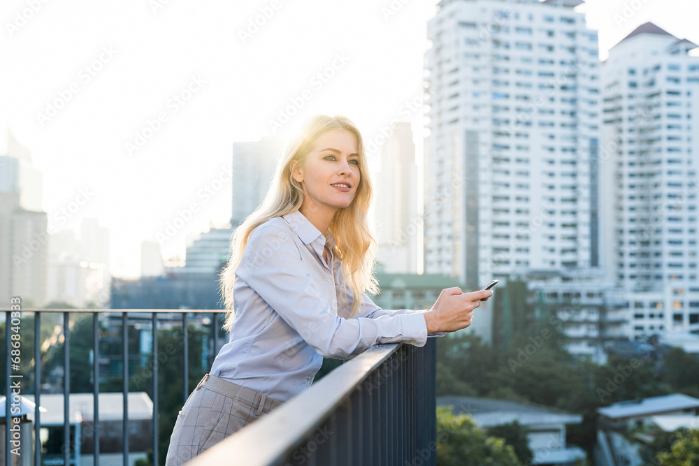 Blonde smiling business woman leaning onto handrail holding smartphone on city rooftop