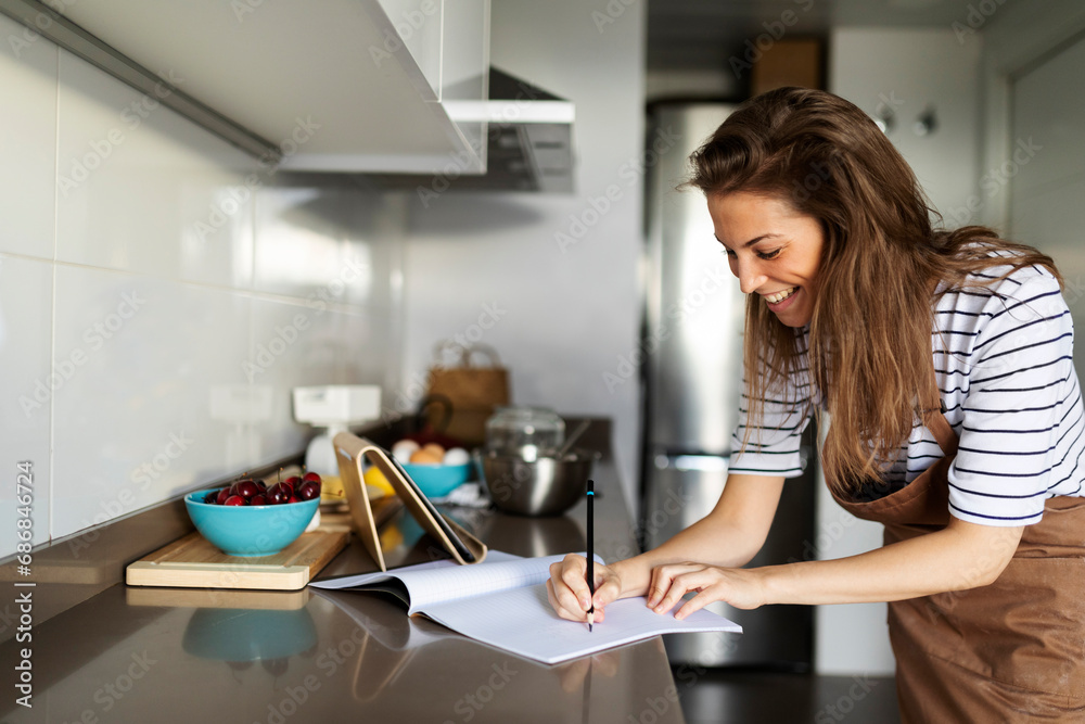 Smiling young woman writing recipe in book while watching video over digital tablet at home Smiling young woman writing recipe in book while watching video over digital tablet at home