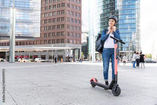 Woman with e-scooter in the city putting on helmet, Berlin, Germany photo