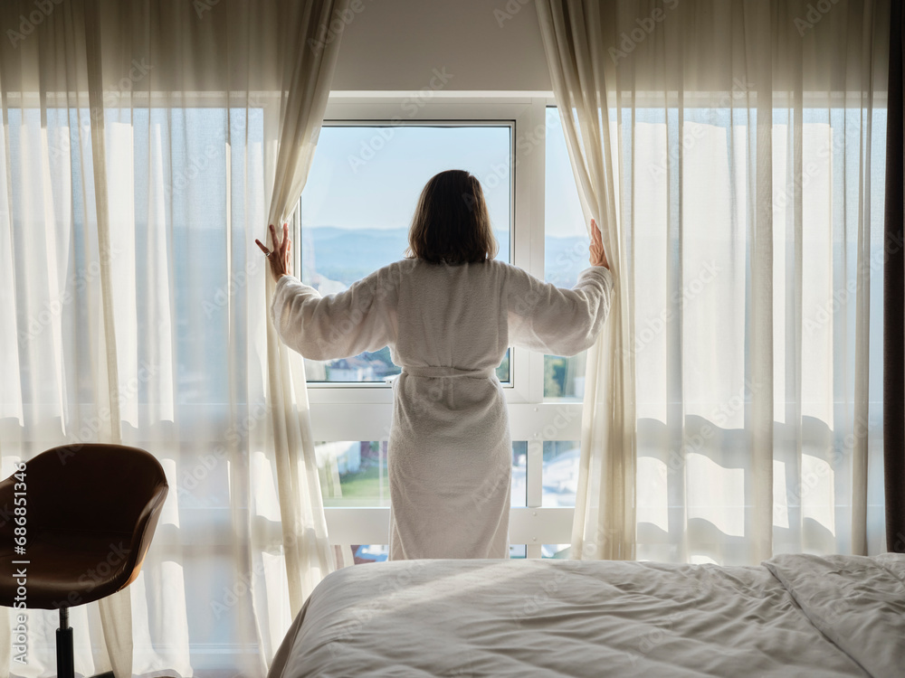 Woman opening white curtain while looking through window at luxury hotel room Woman opening white curtain while looking through window at luxury hotel room