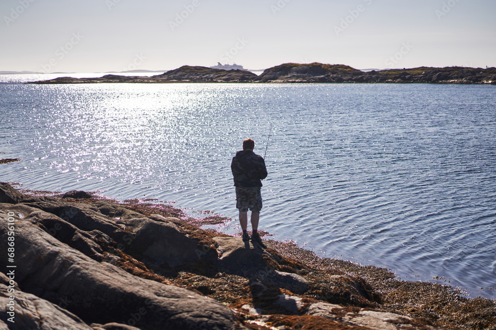 The man with fishing rod spinning the cod fish with soft lures and pilkers on the rocky shore of Norwegian fjord during sunny summer evening. Fishing is popular hobby in all scandinavian countries.