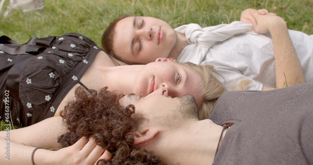 Girl positioned between two guys, tenderly kissing one of them. Love triangles as intricacies of human emotions. Lifestyle portraits of students after a lecture, young people lying on the grass. 