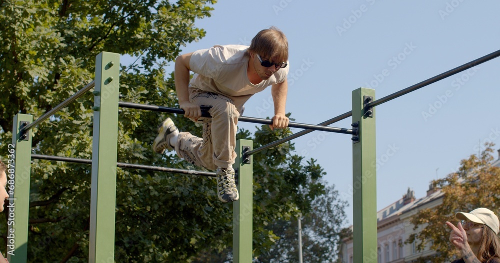 Fototapeta premium Young man performs tricks on horizontal bar in an urban workout area. Physical fitness and exercise outdoor.