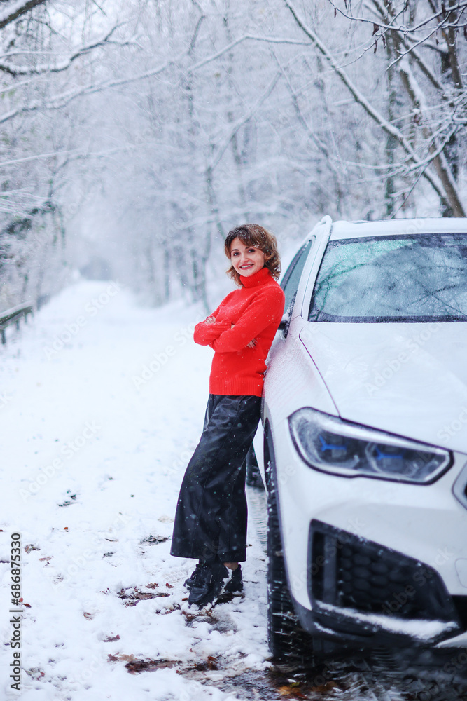 Pretty girl in warm clothes standing in the winter wood while leans on the car.