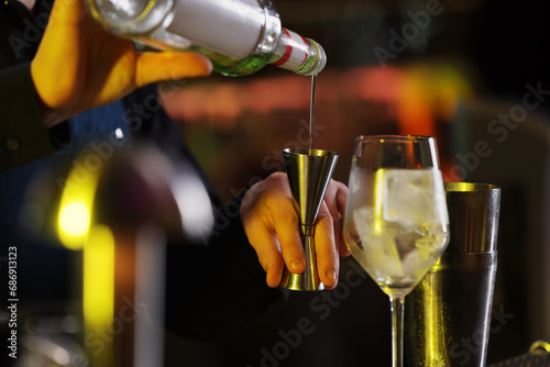 Cocktail making. Bartender pouring alcohol from bottle into jigger on blurred background, closeup photo