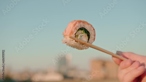 Close-up of man holding with chopsticks a delicious fresh roll of sushi on a summer terrace against the background of the sky