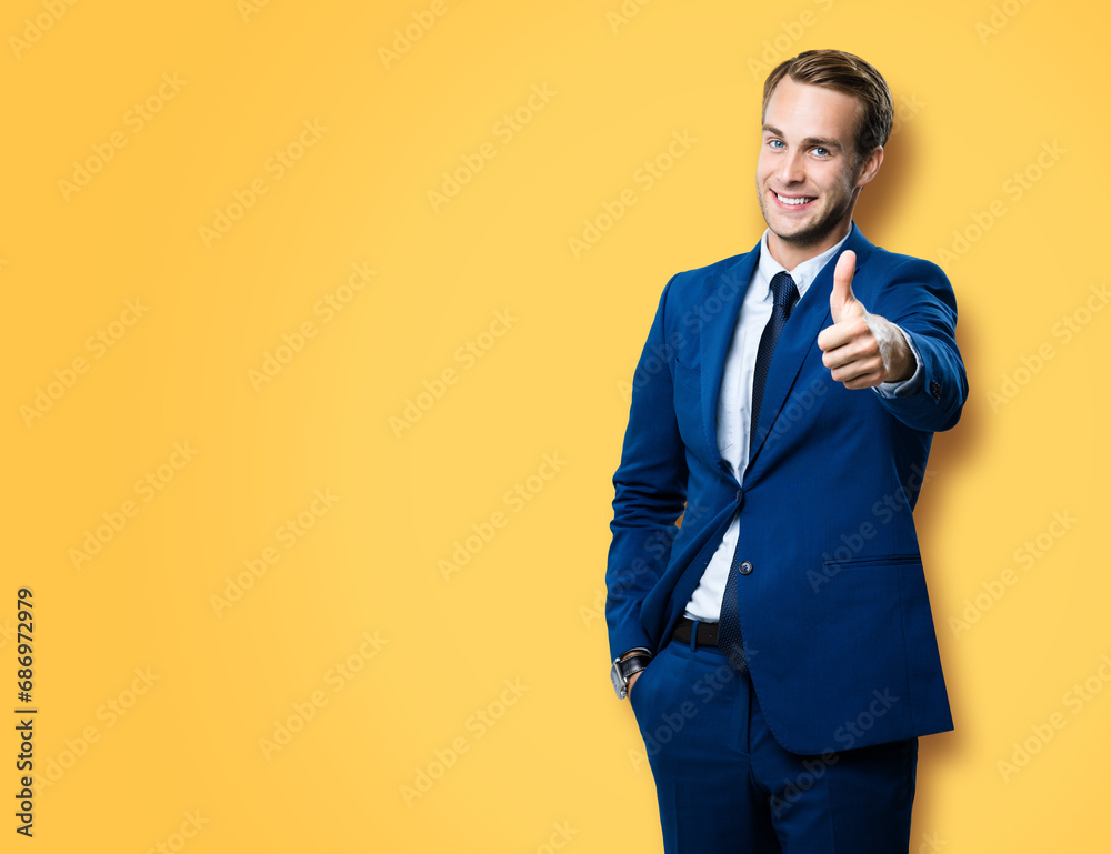 Excited businessman showing thumb up like sign gesture, in blue confident style suit, against vivid yellow background. Handsome happy man. Copy space for slogan or text. Excited businessman showing thumb up like sign gesture, in blue confident style suit, against vivid yellow background. Handsome happy man. Copy space for slogan or text.