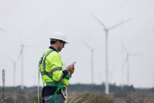Male engineer working with plan maintenance of wind turbines at windmill field farm. Male engineer using mobile phone monitoring wind turbine system at wind turbines farm