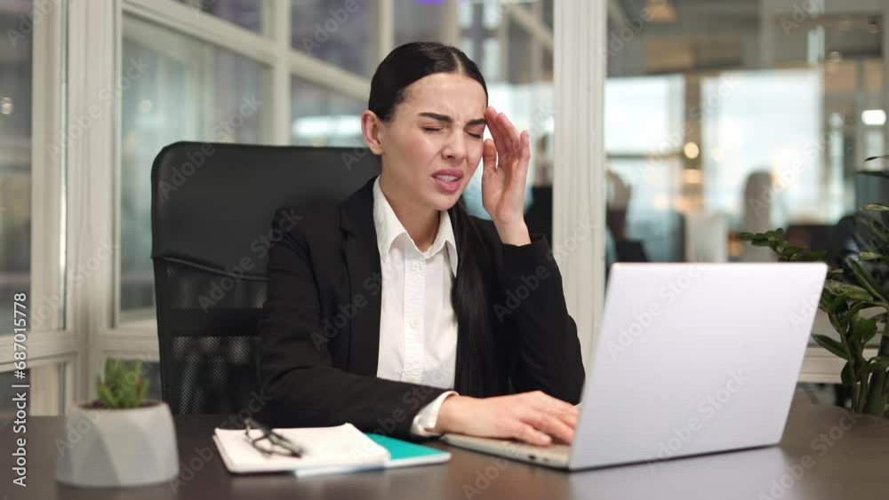 Female office worker sitting and typing on modern laptop keyboard and feeling headache from working with device. Sad brunette lady in suit putting hand to temple and trying ease pain with massage