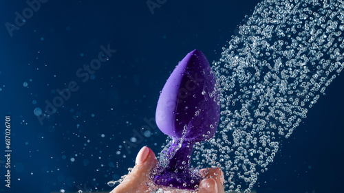Woman washing purple anal plug under shower on blue background. 