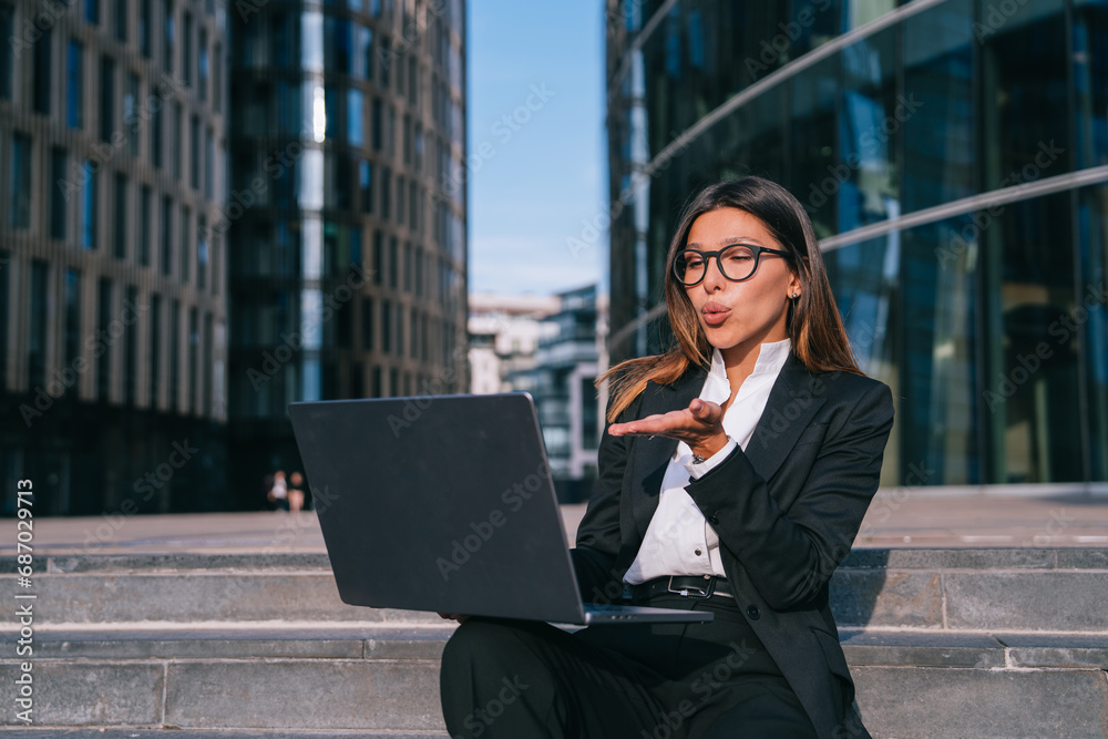 Satisfied young female entrepreneur in business suit sitting on stairs makes video call sends air kiss, deal. Hispanic businesswoman outside, chatting with husband during break of work. Satisfied young female entrepreneur in business suit sitting on stairs makes video call sends air kiss, deal. Hispanic businesswoman outside, chatting with husband during break of work.