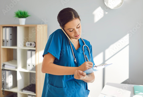 Portrait of a young female doctor or woman nurse standing in office talking by mobile phone and writing notes to clipboard notepad in medical clinic. Medicine and health care concept. photo