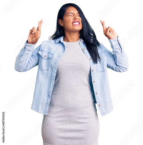 Hispanic woman with long hair wearing casual denim jacket gesturing finger crossed smiling with hope and eyes closed. luck and superstitious concept.