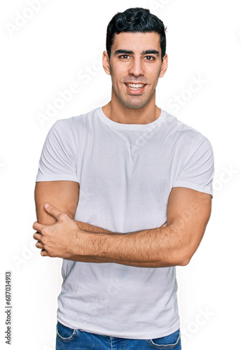 Handsome hispanic man wearing casual white t shirt happy face smiling with crossed arms looking at the camera. positive person.