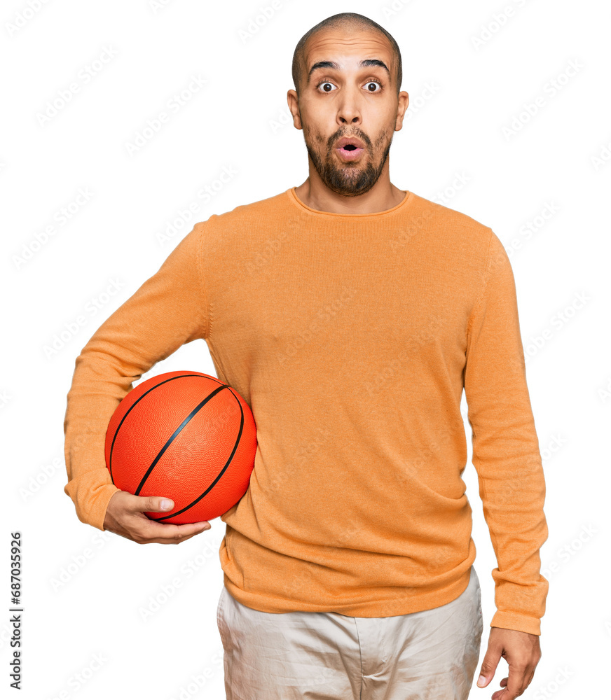 Hispanic adult man holding basketball ball scared and amazed with open mouth for surprise, disbelief face