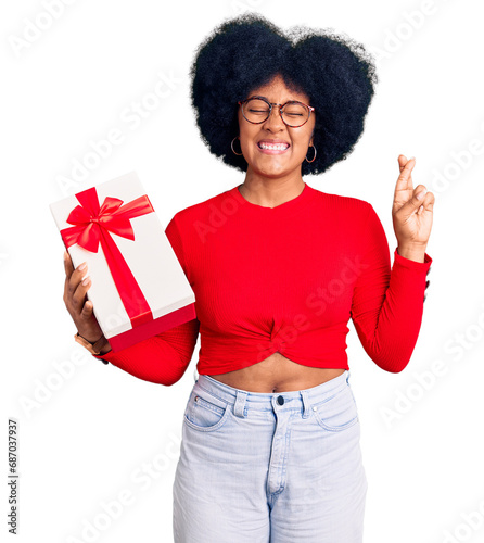 Young african american girl holding gift gesturing finger crossed smiling with hope and eyes closed. luck and superstitious concept.