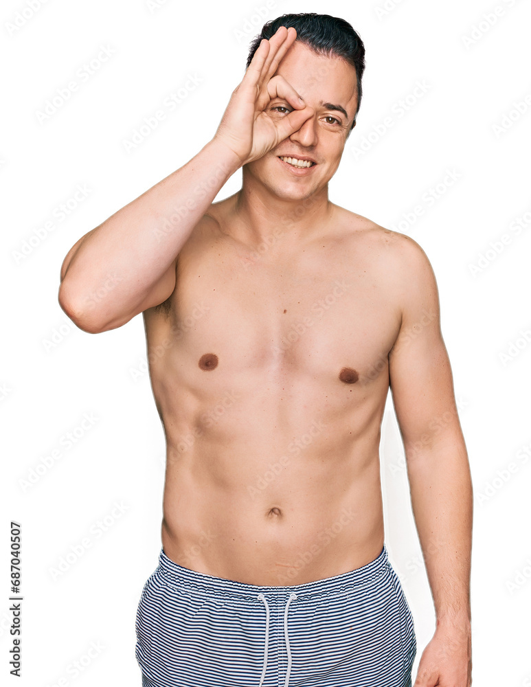 Handsome young man wearing swimwear shirtless doing ok gesture with hand smiling, eye looking through fingers with happy face. Handsome young man wearing swimwear shirtless doing ok gesture with hand smiling, eye looking through fingers with happy face.