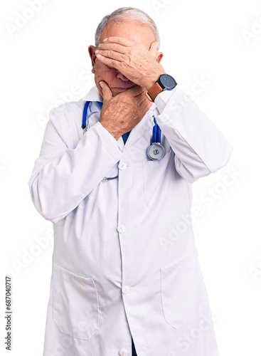 Senior handsome grey-haired man wearing doctor coat and stethoscope covering eyes and mouth with hands, surprised and shocked. hiding emotion