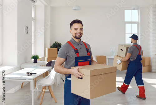 Young happy smiling employee of moving service in overall standing in the living room of new house holding cardboard box and looking cheerful at camera with other mover on background.