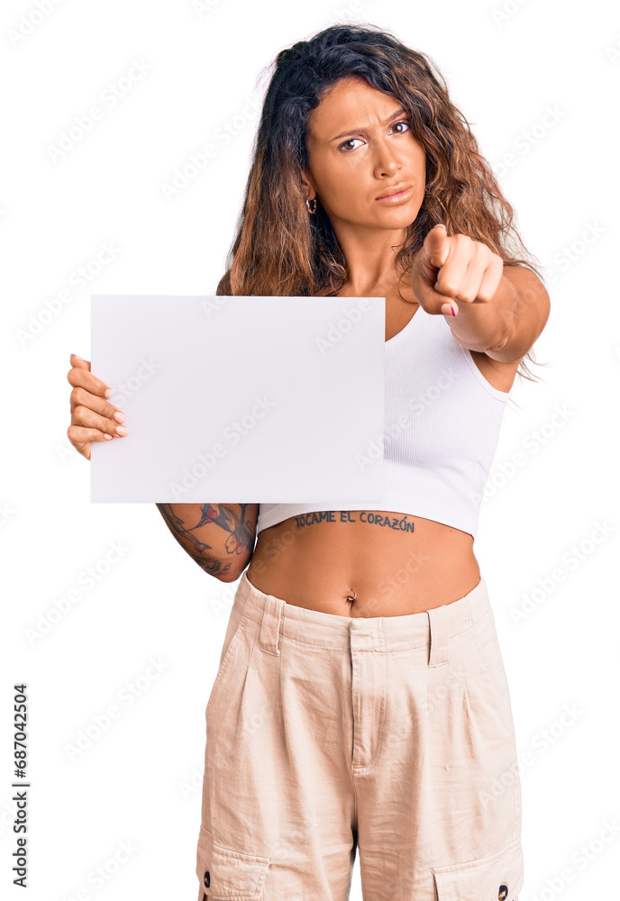 Young hispanic woman with tattoo holding blank empty paper pointing with finger to the camera and to you, confident gesture looking serious