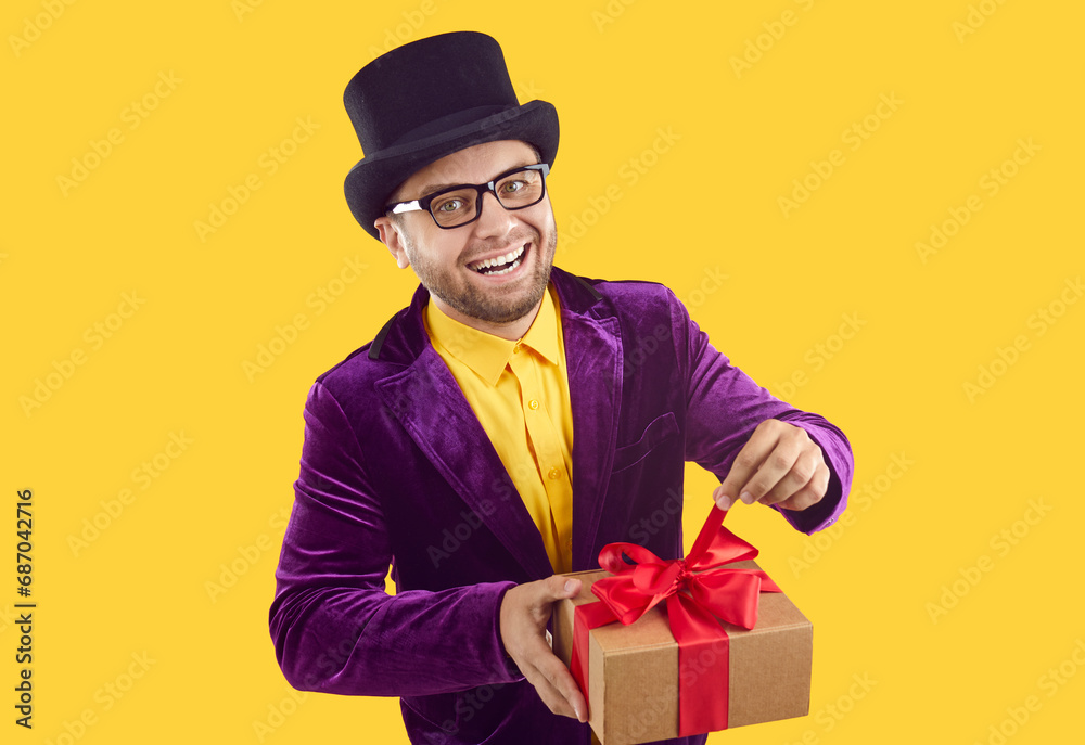 Portrait of funny man with Christmas present. Man in tophat and purple velvet suit standing isolated on yellow background, untying red bow on gift box, smiling, looking at camera with happy expression Portrait of funny man with Christmas present. Man in tophat and purple velvet suit standing isolated on yellow background, untying red bow on gift box, smiling, looking at camera with happy expression
