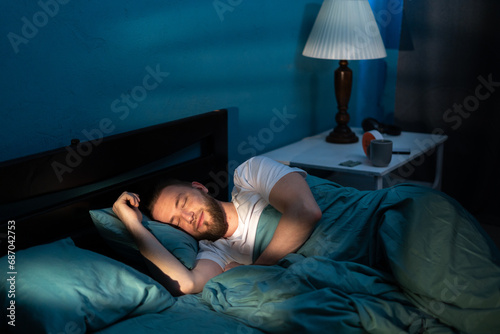 Handsome caucasian man sleeping cozily on a bed in his bedroom at night with blue nightly colors in room photo