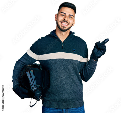 Young hispanic man with beard holding motorcycle helmet smiling happy pointing with hand and finger to the side