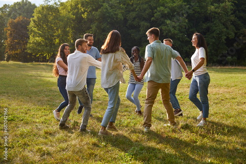 Group of cheerful joyful friends having fun in beautiful summer park. Happy young diverse multiethnic people enjoying summertime, playing silly games and dancing round dance on green lawn all together