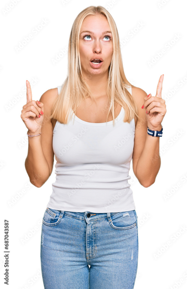 Young blonde girl wearing casual style with sleeveless shirt amazed and surprised looking up and pointing with fingers and raised arms. Young blonde girl wearing casual style with sleeveless shirt amazed and surprised looking up and pointing with fingers and raised arms.