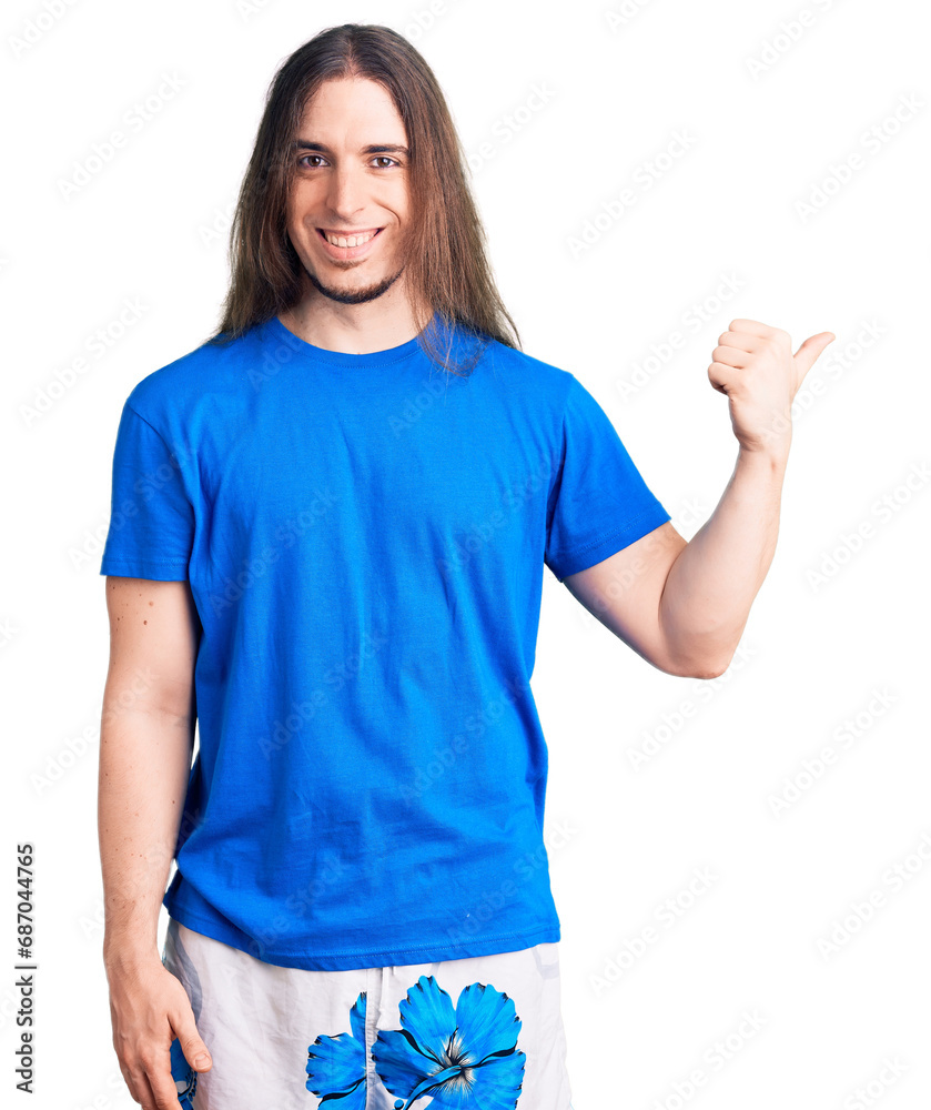 Young adult man with long hair wearing swimwear smiling with happy face looking and pointing to the side with thumb up. Young adult man with long hair wearing swimwear smiling with happy face looking and pointing to the side with thumb up.