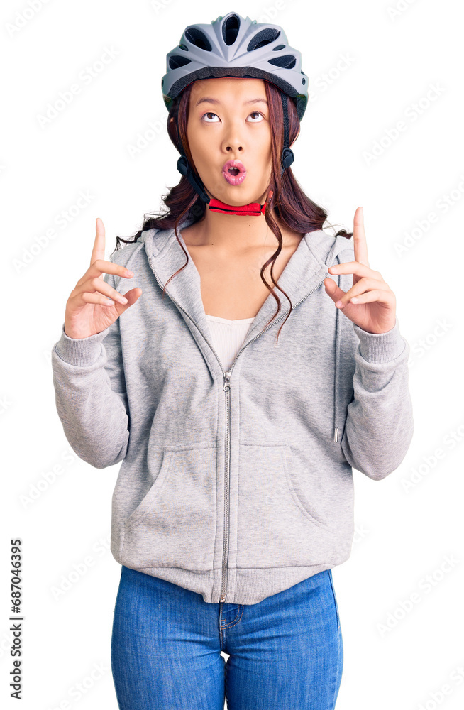 Young beautiful chinese girl wearing bike helmet amazed and surprised looking up and pointing with fingers and raised arms. Young beautiful chinese girl wearing bike helmet amazed and surprised looking up and pointing with fingers and raised arms.