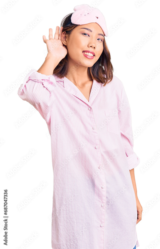 Young beautiful chinese girl wearing sleep mask and pajama smiling with hand over ear listening an hearing to rumor or gossip. deafness concept. Young beautiful chinese girl wearing sleep mask and pajama smiling with hand over ear listening an hearing to rumor or gossip. deafness concept.