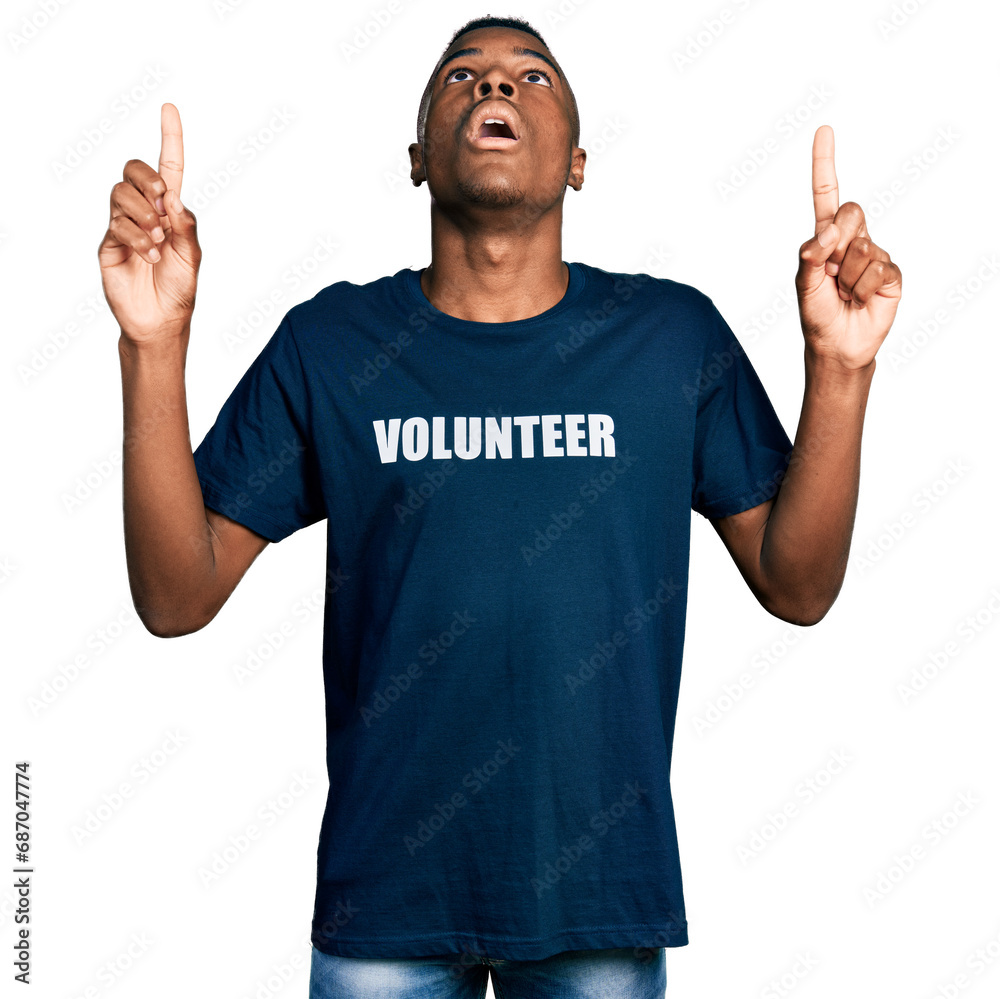 Young african american man wearing volunteer t shirt amazed and surprised looking up and pointing with fingers and raised arms. Young african american man wearing volunteer t shirt amazed and surprised looking up and pointing with fingers and raised arms.