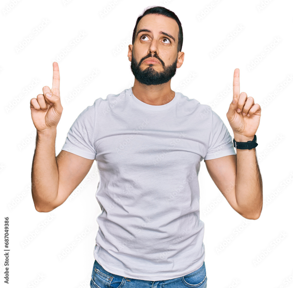 Young man with beard wearing casual white t shirt pointing up looking sad and upset, indicating direction with fingers, unhappy and depressed. Young man with beard wearing casual white t shirt pointing up looking sad and upset, indicating direction with fingers, unhappy and depressed.